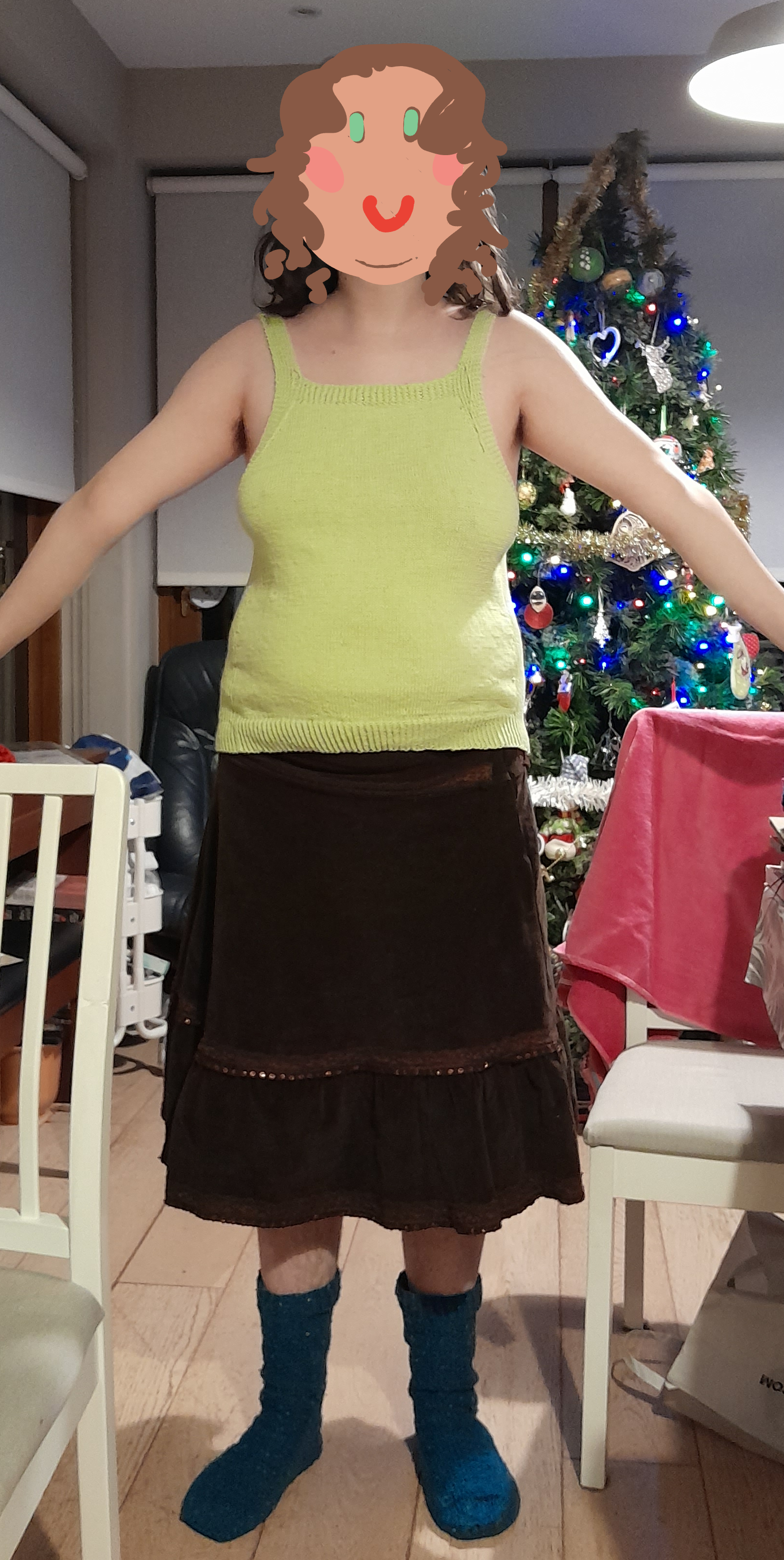 a midsized white person with long hair standing in front of a christmas tree wearing a knitted light green sleeveles top and a brown courdoroy skirt. their face is painted over with a childlike drawing of a white face with green eyes smiling.